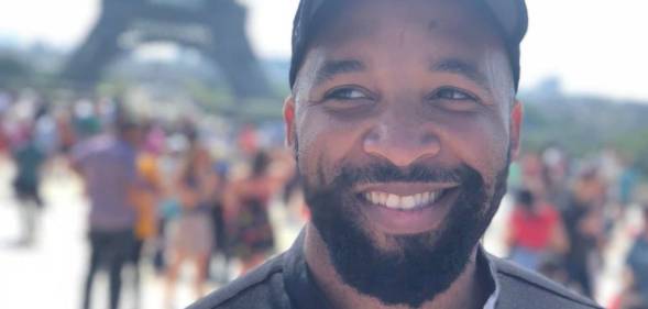Michael Reynolds, a Black LGBTQ+ gamer with a beard, smiles as he stares off screen. He is wearing a black shirt and black cap as he stands outside
