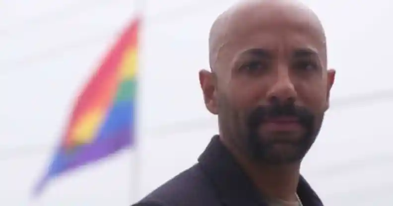 Nas Mohamed looks to the camera in front of an LGBTQ+ Pride flag