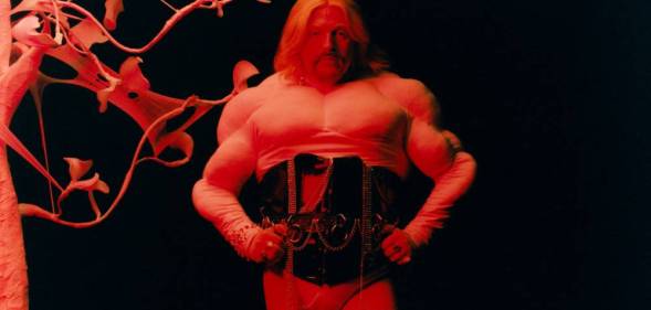 Tom Rasmussen wears a white top and black corset as they stand with their hands on their hips and are lit by a red light