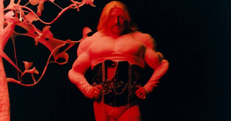 Tom Rasmussen wears a white top and black corset as they stand with their hands on their hips and are lit by a red light