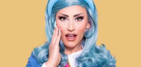 Drag Race UK season three queen Ella Vaday poses for the camera with her hand on her cheek while wearing a blue wig, blue headband, blue jacket, white shirt and pink, black and yellow striped tie