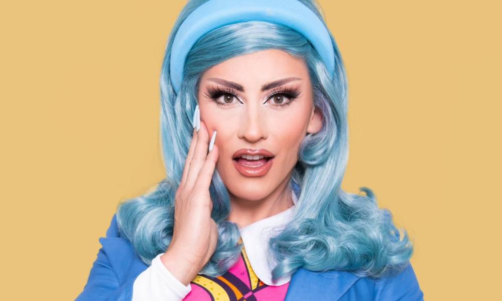 Drag Race UK season three queen Ella Vaday poses for the camera with her hand on her cheek while wearing a blue wig, blue headband, blue jacket, white shirt and pink, black and yellow striped tie
