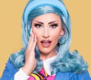 Drag Race UK season three queen Ella Vaday poses for the camera with her hand on her cheek while wearing a blue wig, blue headband, blue jacket, white shirt and pink, black and yellow striped tie