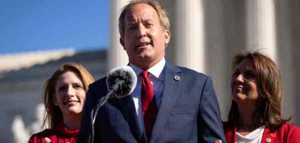 Texas attorney general Ken Paxton wears a white button up shirt, red tie and blue suit jacket as he speaks into a microphone and gestures with both his hands. He is standing in front of two people wearing red outfits