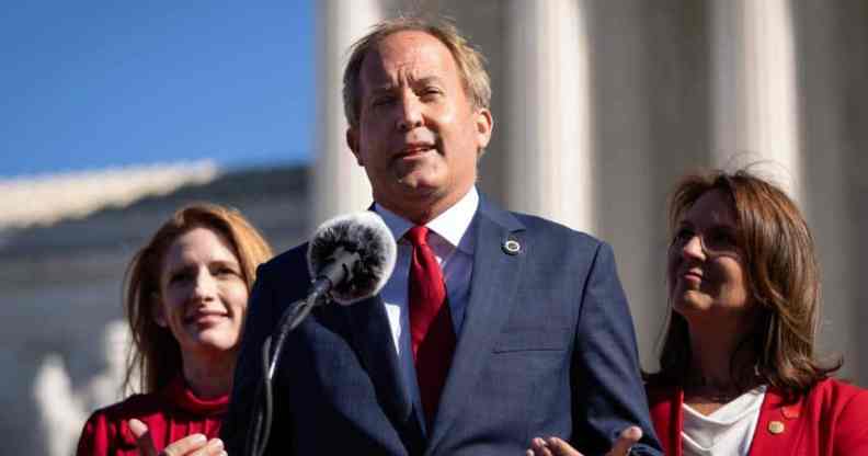 Texas attorney general Ken Paxton wears a white button up shirt, red tie and blue suit jacket as he speaks into a microphone and gestures with both his hands. He is standing in front of two people wearing red outfits