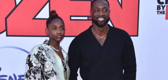 Dwyane Wade wears a black shirt and smiles as he stands next to his daughter Zaya who is wearing a black and white floral patterned top