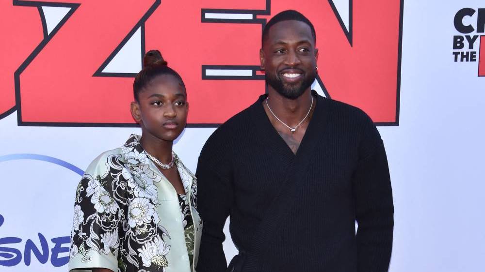 Dwyane Wade wears a black shirt and smiles as he stands next to his daughter Zaya who is wearing a black and white floral patterned top