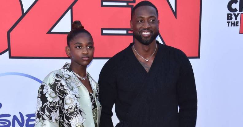 Dwyane Wade wears a black shirt and smiles as he stands next to his daughter Zaya who is wearing a black and white floral patterned top