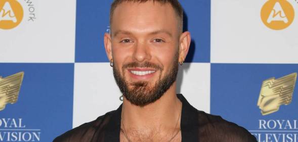 John Whaite smiles at the camera while wearing a black mesh top with a gold necklace as he stands in front of a white and blue checkered background