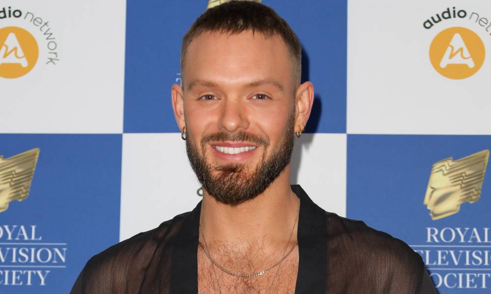 John Whaite smiles at the camera while wearing a black mesh top with a gold necklace as he stands in front of a white and blue checkered background