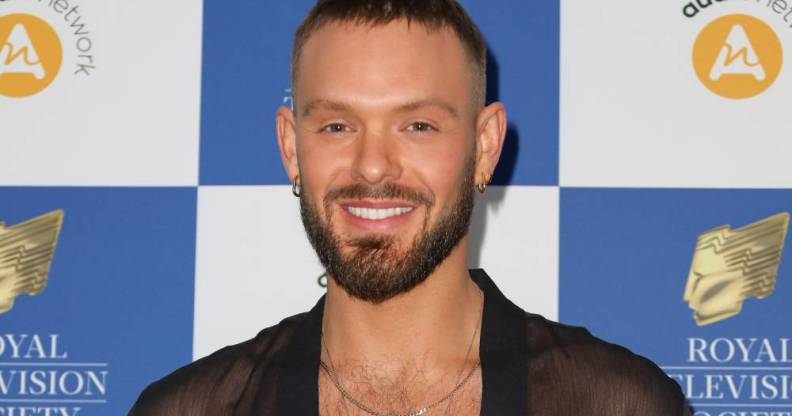 John Whaite smiles at the camera while wearing a black mesh top with a gold necklace as he stands in front of a white and blue checkered background