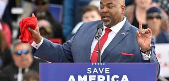 North Carolina lieutenant governor Mark Robinson wears a white button up shirt, red tie, blue suit jacket and red pocket square as he stands at a podium with a sign reading 'Save America' on it. He is gesturing with both his hands in the air and one hand is holding a red towel