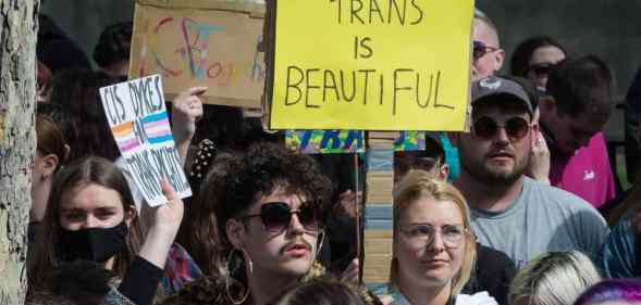Several people gather in a crowd in support of the trans community in the UK. One person holds up a sign reading 'trans is beautiful' in the crowd.