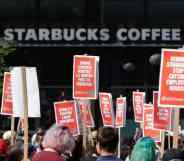 People in a crowd hold up various orange signs with white writing in support of Starbucks unionising with a white Starbucks logo from a store is seen in the background