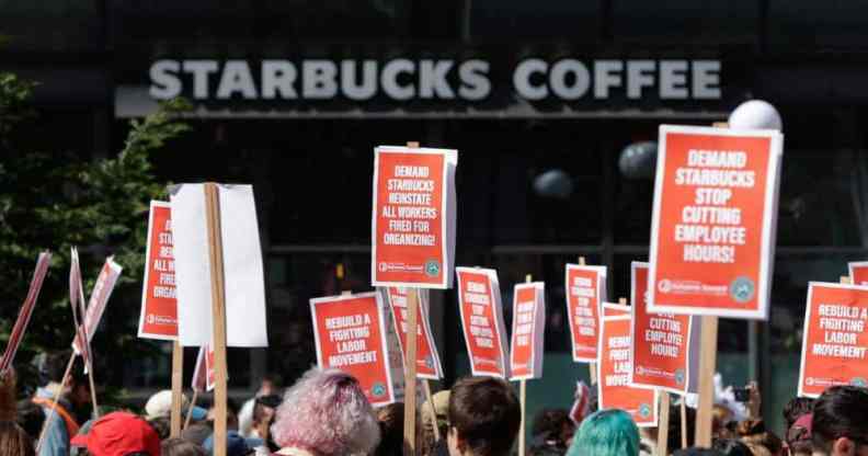 People in a crowd hold up various orange signs with white writing in support of Starbucks unionising with a white Starbucks logo from a store is seen in the background