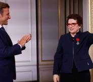 French president Emmanuel Macron wears a dark suit and white button up shirt as he stands sideways and claps for Billie Jean King, who is dressed in a black outfit with a navy blue jacket on top. She has the France's Legion of Honour award pinned to her chest and raises her left arm in the air, and she is wearing a rainbow bracelet around her wrist
