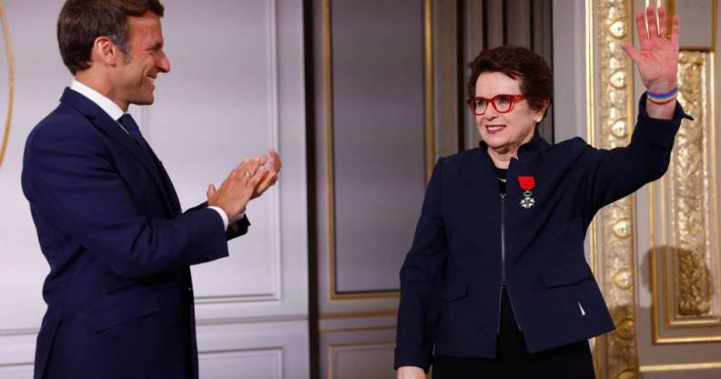 French president Emmanuel Macron wears a dark suit and white button up shirt as he stands sideways and claps for Billie Jean King, who is dressed in a black outfit with a navy blue jacket on top. She has the France's Legion of Honour award pinned to her chest and raises her left arm in the air, and she is wearing a rainbow bracelet around her wrist
