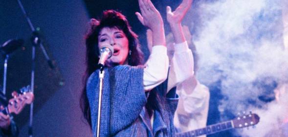 Kate Bush wears a white top and blue long sweater as she sings into a microphone and claps her hands above her head during a live performance