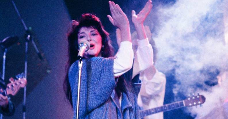 Kate Bush wears a white top and blue long sweater as she sings into a microphone and claps her hands above her head during a live performance