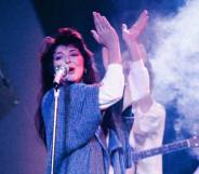 Kate Bush wears a white top and blue long sweater as she sings into a microphone and claps her hands above her head during a live performance