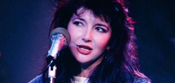 Kate Bush wears a white white shirt, long blue jumped as she sings into a microphone