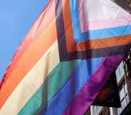 The progressive Pride flag flies outside a building, against a bright blue sky.