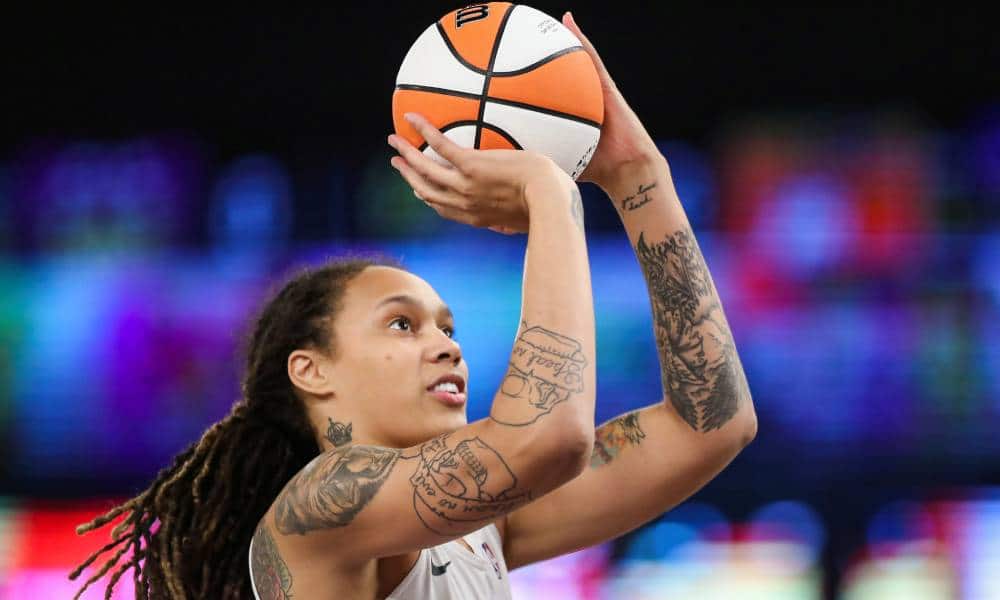 Brittney Griner wears a white jersey as she holds up a white and orange basketball as she shoots it towards a hoop. Her hair is styled in locks and she has tattoos on her arms