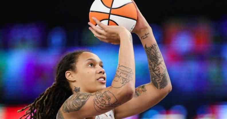 Brittney Griner wears a white jersey as she holds up a white and orange basketball as she shoots it towards a hoop. Her hair is styled in locks and she has tattoos on her arms