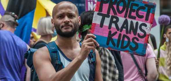 A person holds up a sign coloured in blue, pink and white like the trans Pride flag which reads 'Protect trans kids'