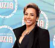 Dame Kelly Holmes wears a black top and black jacket as she smiles at the camera and stands in front of a blue background