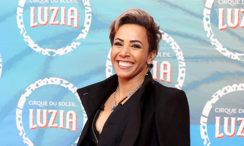 Dame Kelly Holmes wears a black top and black jacket as she smiles at the camera and stands in front of a blue background