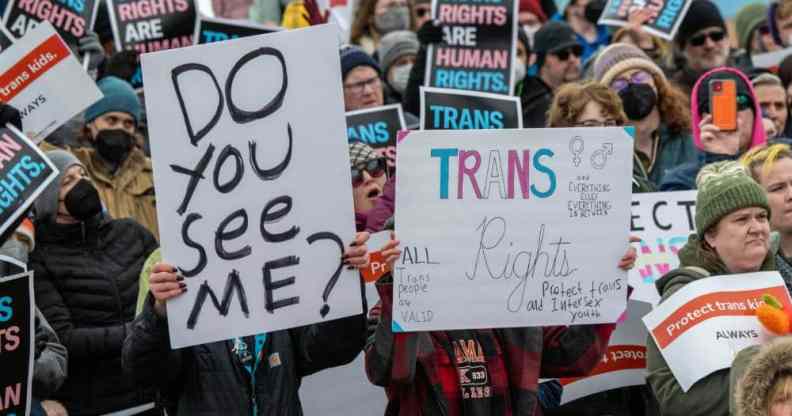 Several people protest in support of trans rights and condemning trans sports bans bills popping up across the US. One person carries a sign reading 'Do you see me?' while the person nexts to them has a sign reading 'Trans rights'. In the background, several people are holding 'Trans rights are human rights' jackets