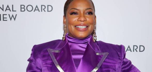 Aunjanue Ellis smiles at the camera while wearing a bright purple turtleneck top and matching jacket with silver lining on the lapel. She is wearing gold dangle earrings and pinky-purple eyeshadow to match her clothing