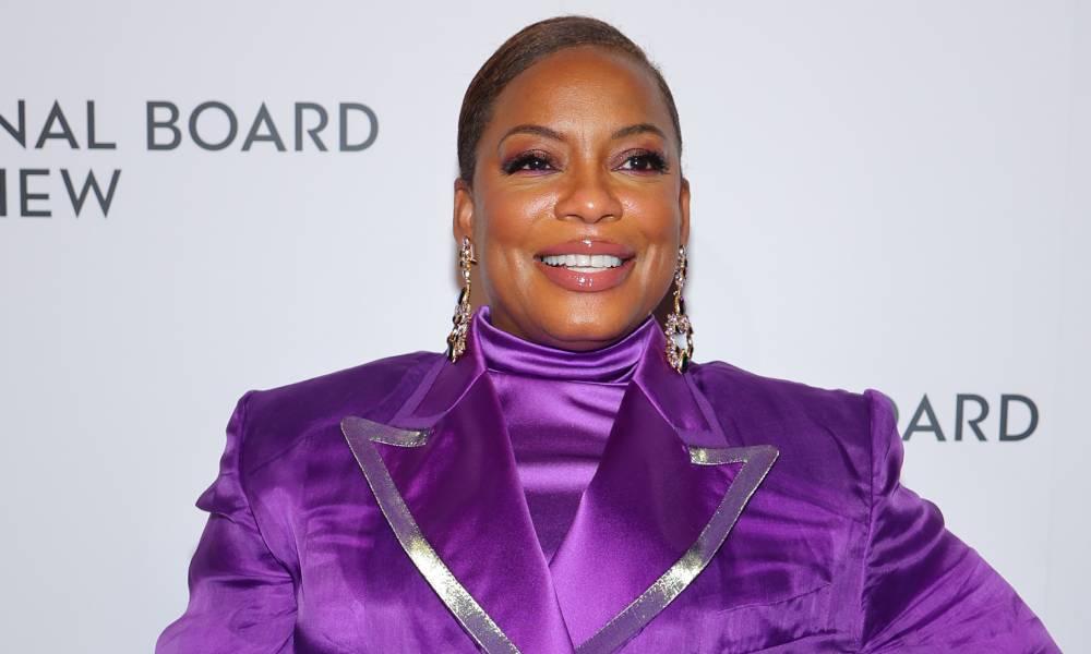 Aunjanue Ellis smiles at the camera while wearing a bright purple turtleneck top and matching jacket with silver lining on the lapel. She is wearing gold dangle earrings and pinky-purple eyeshadow to match her clothing