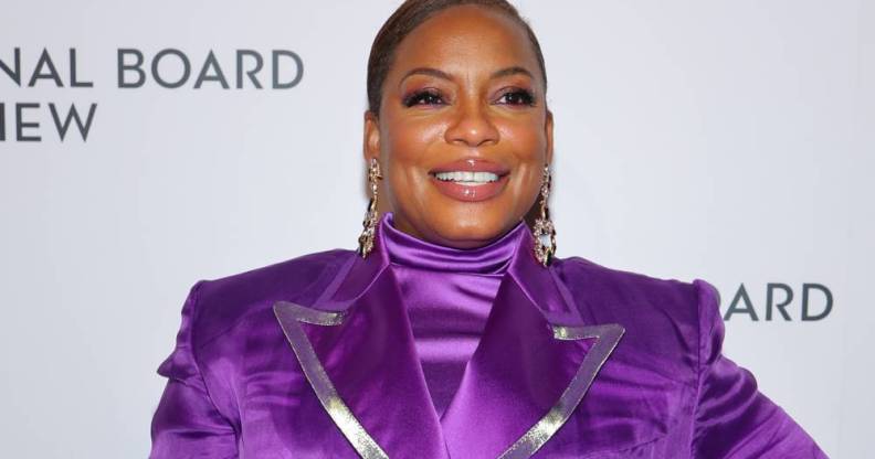 Aunjanue Ellis smiles at the camera while wearing a bright purple turtleneck top and matching jacket with silver lining on the lapel. She is wearing gold dangle earrings and pinky-purple eyeshadow to match her clothing
