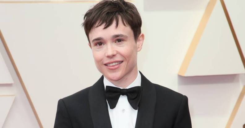 Elliot Page smiles for the camera while wearing a bow tie, white button up shirt and black suit jacket and standing in front of a pale off-white background