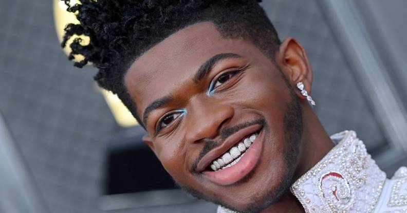 Lil Nas X smiles at the camera showing off decorative sparkly grills on his teeth. He has light blue eye shadow at the inner corner of his eyes and is wearing a while outfit with sparkly details on the collar and a matching dangly earring in his ear