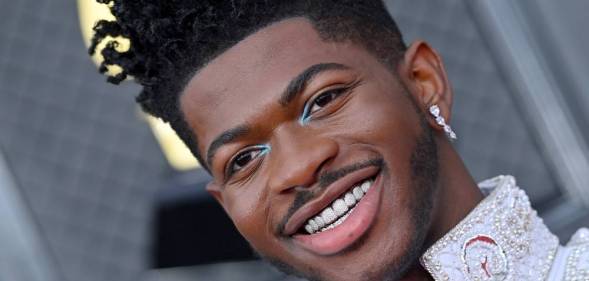 Lil Nas X smiles at the camera showing off decorative sparkly grills on his teeth. He has light blue eye shadow at the inner corner of his eyes and is wearing a while outfit with sparkly details on the collar and a matching dangly earring in his ear