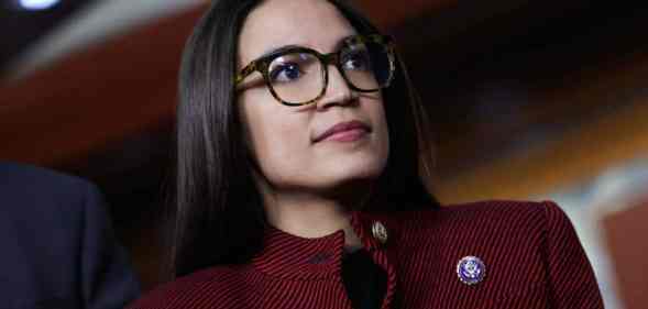 Alexandria Ocasio-Cortez, known as AOC, wears a red and black striped jacket with a dark top underneath with a little pin as she stares at something off camera