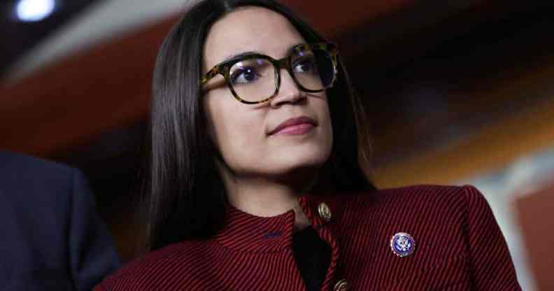 Alexandria Ocasio-Cortez, known as AOC, wears a red and black striped jacket with a dark top underneath with a little pin as she stares at something off camera