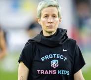 Megan Rapinoe stands on a soccer field while wearing a dark, short-sleeved t-shirt that has the words 'protect trans kids' written on it in blue and pink lettering