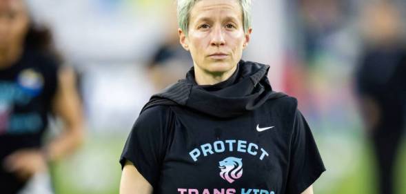 Megan Rapinoe stands on a soccer field while wearing a dark, short-sleeved t-shirt that has the words 'protect trans kids' written on it in blue and pink lettering
