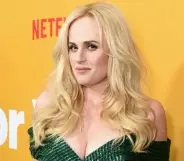 Rebel Wilson wears a green dress with her blonde hair down as she stands in front of a yellow background with the red Netflix logo in the background