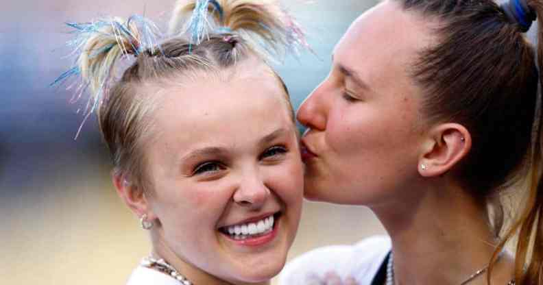 JoJo Siwa smiles at the camera while her hair is in small pig tails with blue sparkly bits sticking out of it while Kylie Prew kisses her cheek