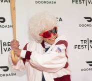 Maybelle Blair, an original All-American Girls Professional Baseball League player, wears a white and red coat with dark red rimmed glasses and she poses for the cameras with a baseball bat as if she is ready to hit a ball