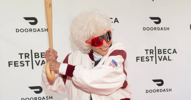 Maybelle Blair, an original All-American Girls Professional Baseball League player, wears a white and red coat with dark red rimmed glasses and she poses for the cameras with a baseball bat as if she is ready to hit a ball