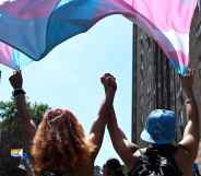 Two trans people hold hands while marching under a trans pride flag