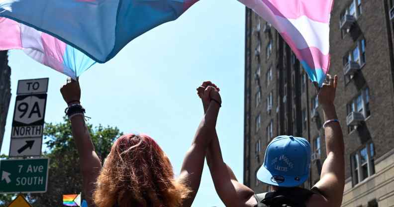 Two trans people hold hands while marching under a trans pride flag