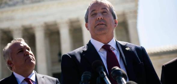 Texas attorney general Ken Paxton wears a white button up shirt, red tie and black suit jacket as he speaks into a microphone