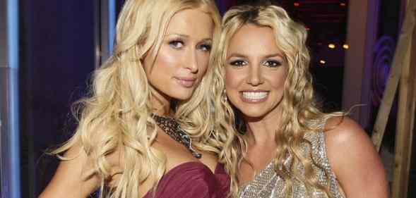 Paris Hilton wears a strapless red dress with a large necklace with her blonde hair down in waves as she stands next to Britney Spears. Spears smiles as she wears a silver, sparkly dress with her blonde hair down in waves
